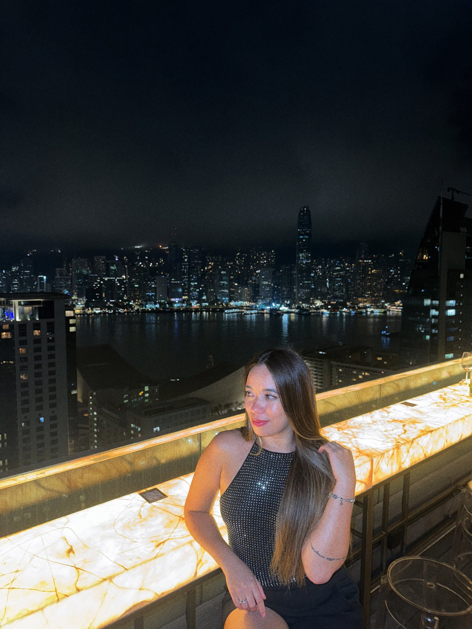 Denise num rooftop com a baía de Hong Kong ao fundo, de noite